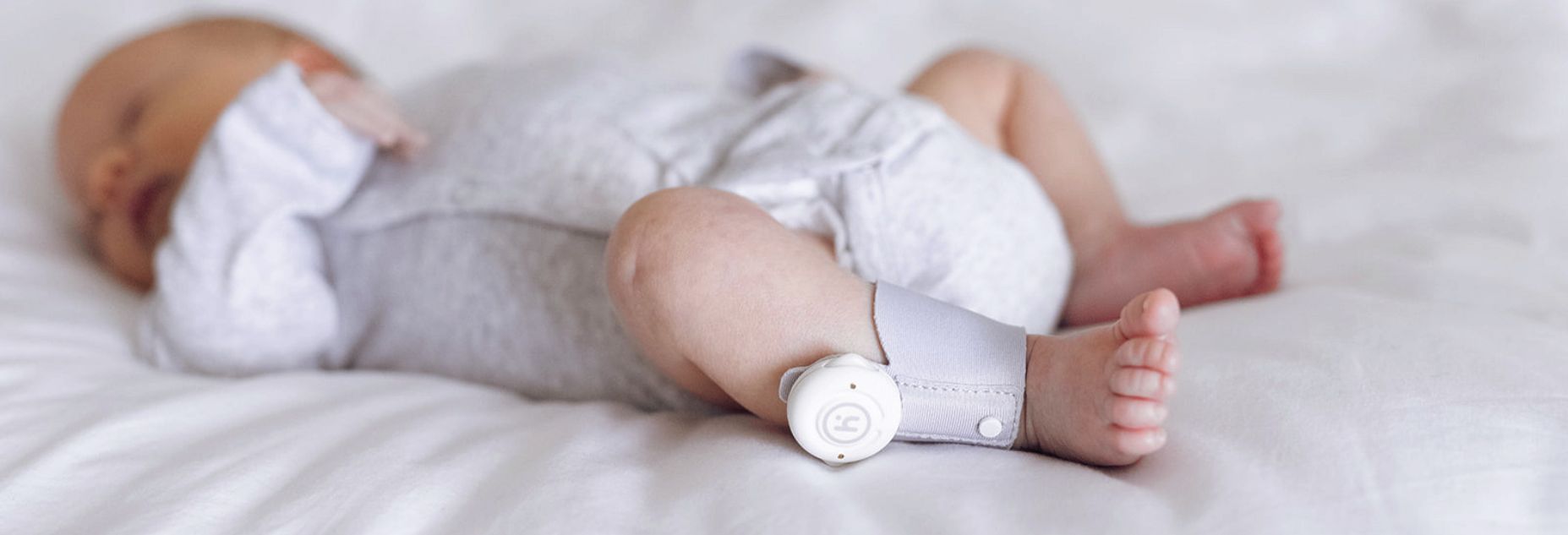 Sleeping baby wearing a smart monitor on their ankle, lying on a soft white bed.